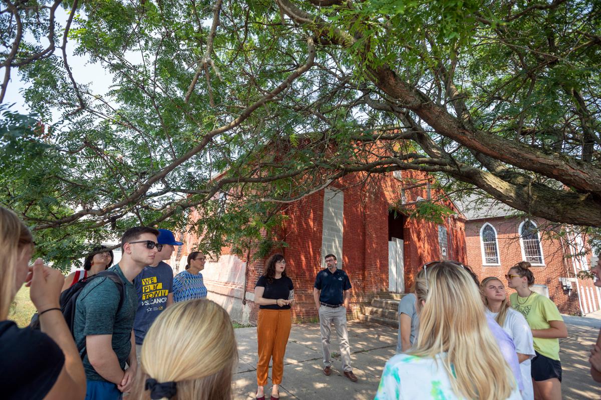 A group of people are gathered under a tree near a brick building, listening to someone speak.