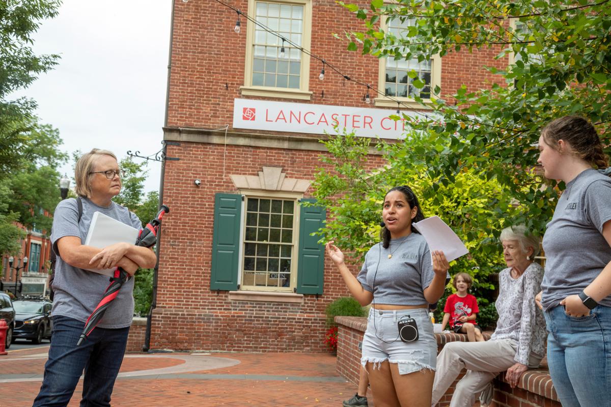 A young woman speaks passionately to a group outside a brick building labeled "Lancaster City."