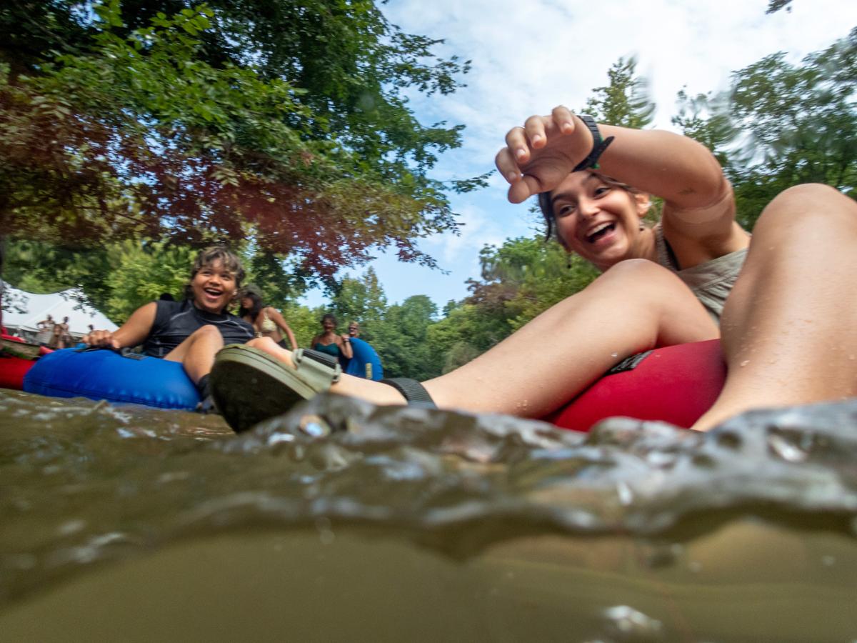 Two people are joyfully tubing down a river, with trees and a blue sky in the background.