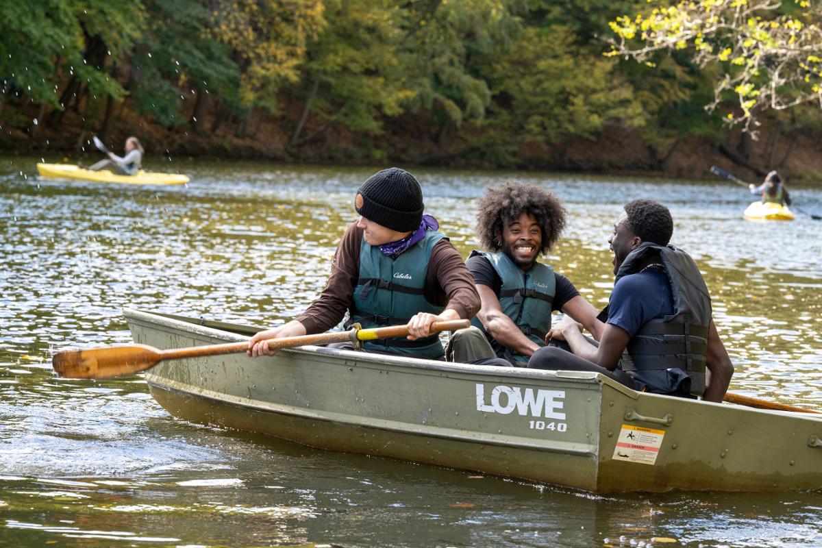 Three students enjoying a boat ride on a river, with others kayaking in the background.