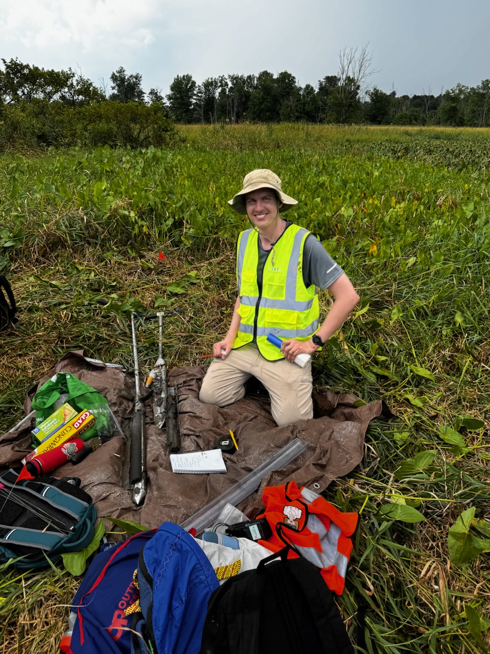 a photo of Adam Benfield kneeling in a meadow while wearing a florescent yellow safety vest