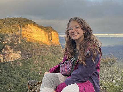 A person sits outside overlooking cliffs at sunset.