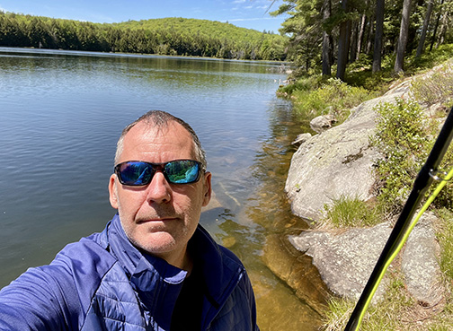 Photo of Chris Williams by a lake holding a fishing rod. 