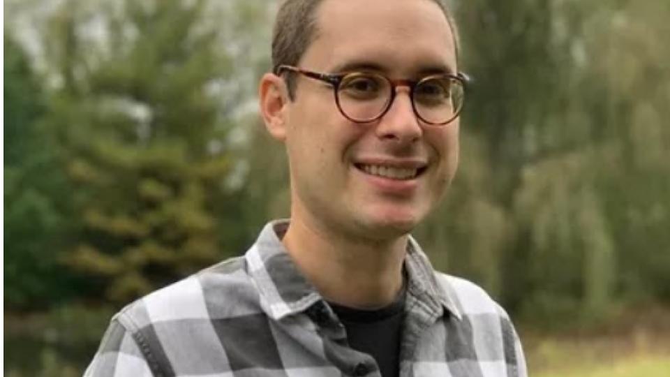 A man with glasses stands where a meadow meets a forest
