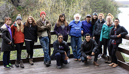 A white haired and white bearded man stands with students on a deck over a lake